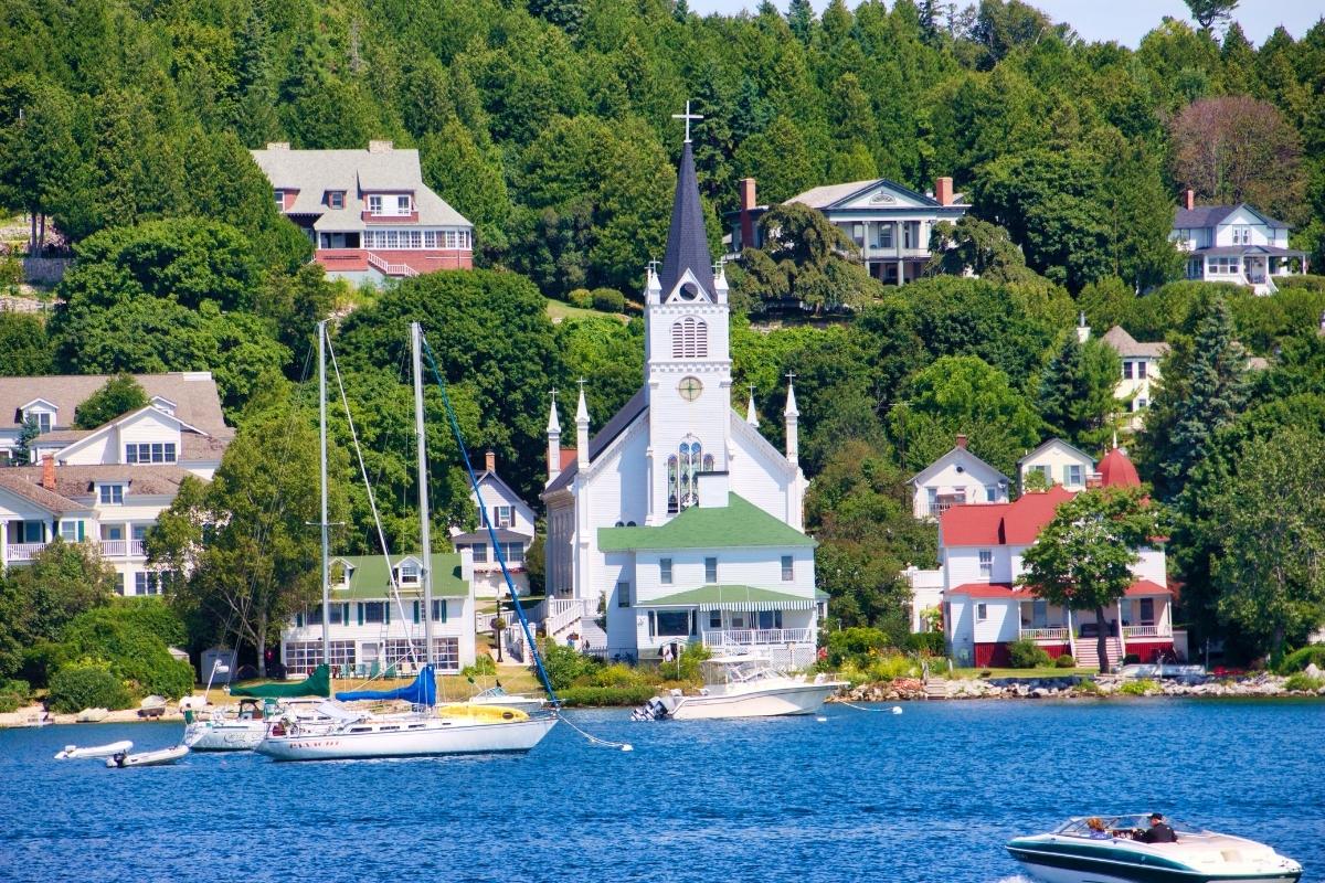 White church and lake houses surrounded by green trees, next to blue water (with boats) on Mackinac Island