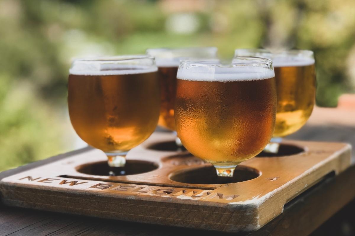 Four golden brown beers in cold glasses with condensation on outside, set in a brown tray on a brown wooden tabletop outdoors with blurry green trees in the background