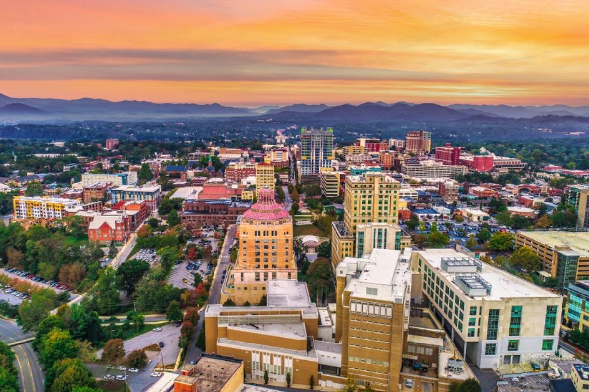 Aerial view of downtown Asheville with buildings, trees, roads, cars, purple mountains and orange-tinged sky in the background