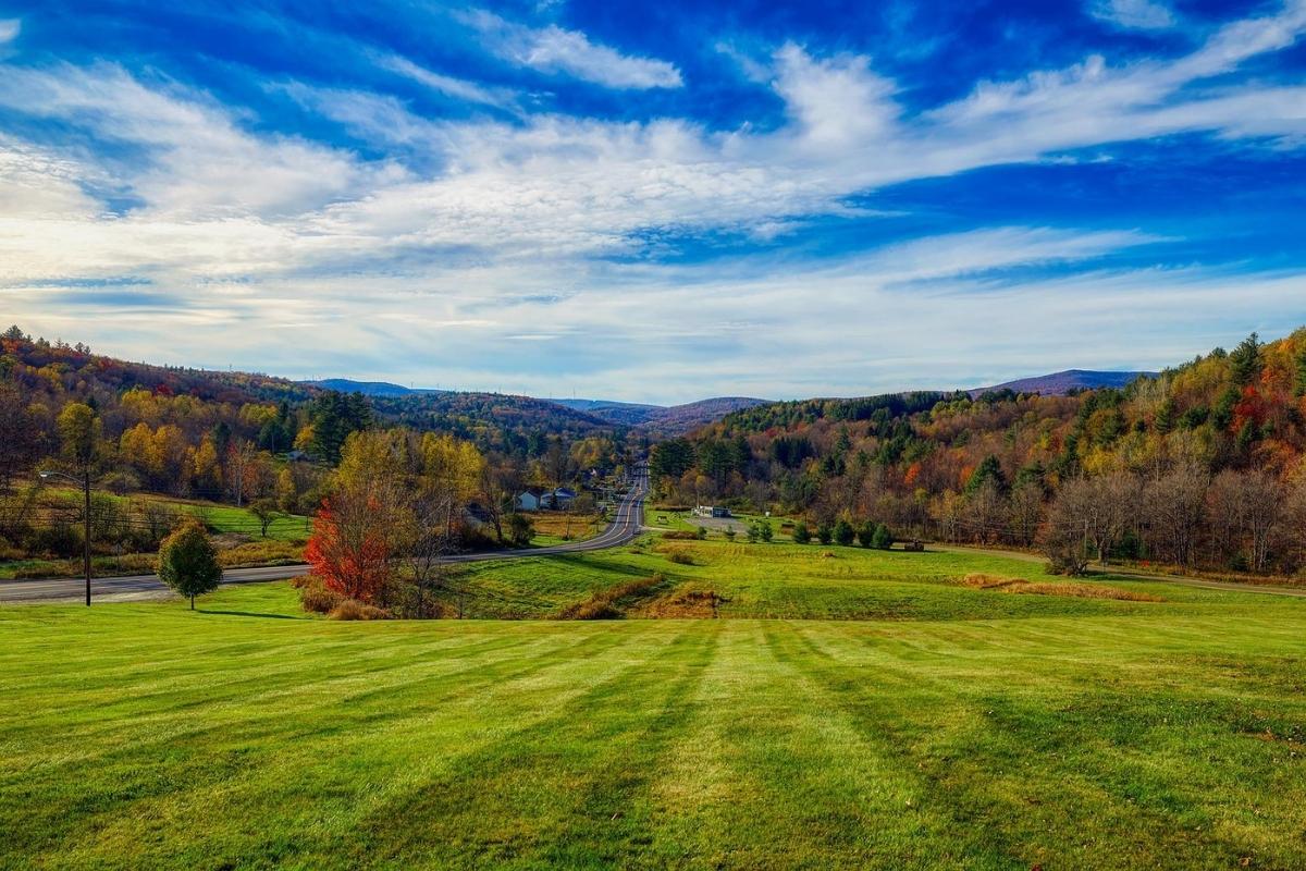 Open green field next to a two-lane road, surrounded by trees with red, orange, and gold fall colors, mountains and small buildings in the distance, and blue sky/white clouds overhead.