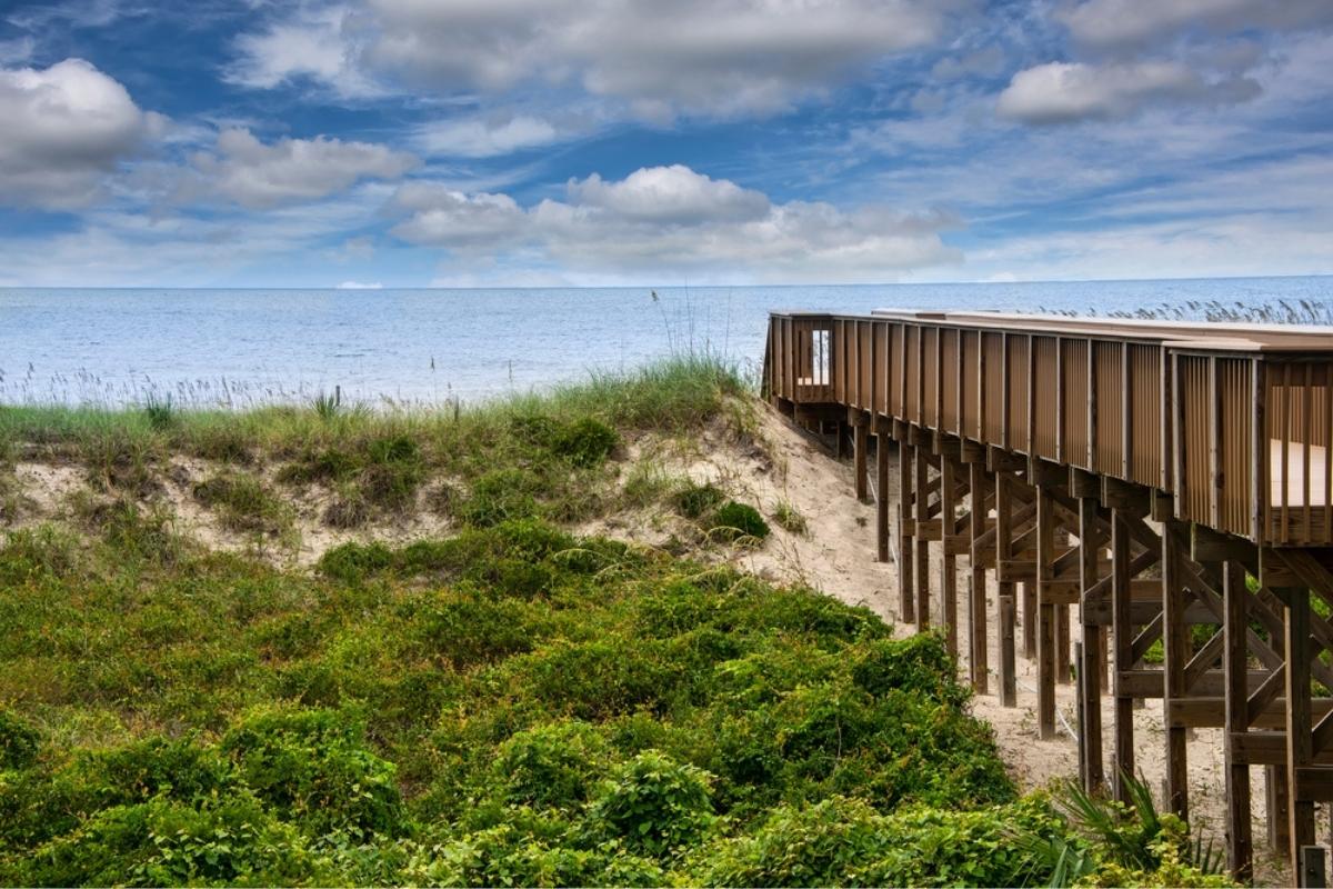 Grass-covered sand dunes next to a brown wooden boardwalk with railings, leading to a beach on Amelia Island. The water is a light shade of blue with only ripples of waves and the sky overhead is a deep blue with fluffy white clouds.