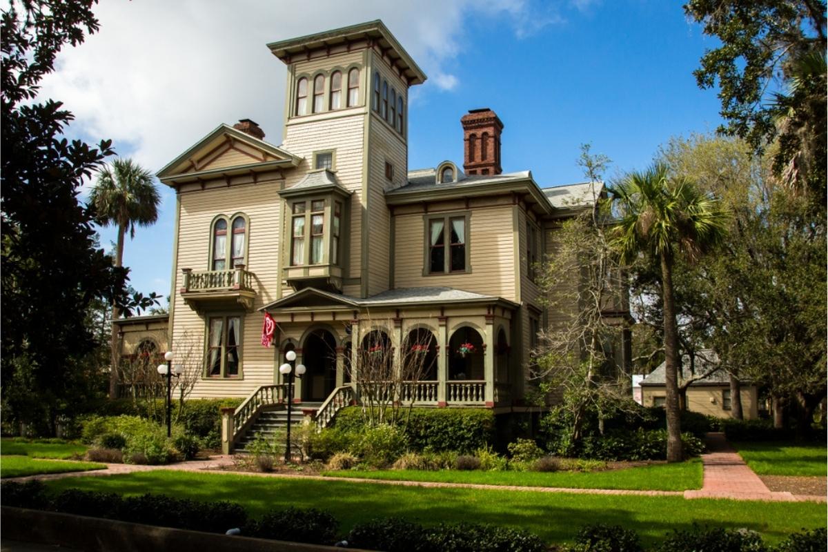 Historic two story wooden home set on a large green lot with lots of trees and landscaping on Amelia Island. there's brick walkway leading from the front and side up to a covered front porch with red flag and hanging flower baskets. The sky overhead is blue with white clouds.