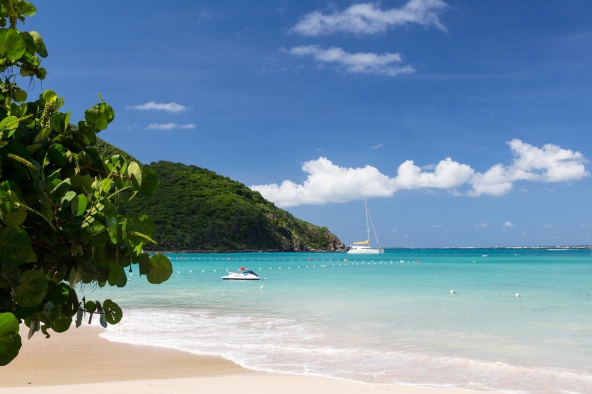 View of Anse Marcel beach in St. Martin, with small stretch of golden sand, blue green water, a green covered hill in the background, and green leaves on a tree in the foreground. The sky is dark blue with white clouds. Part of the water is roped off with white floats and rope, and there is a sailboat (with sails down) and a jet ski empty in the water.