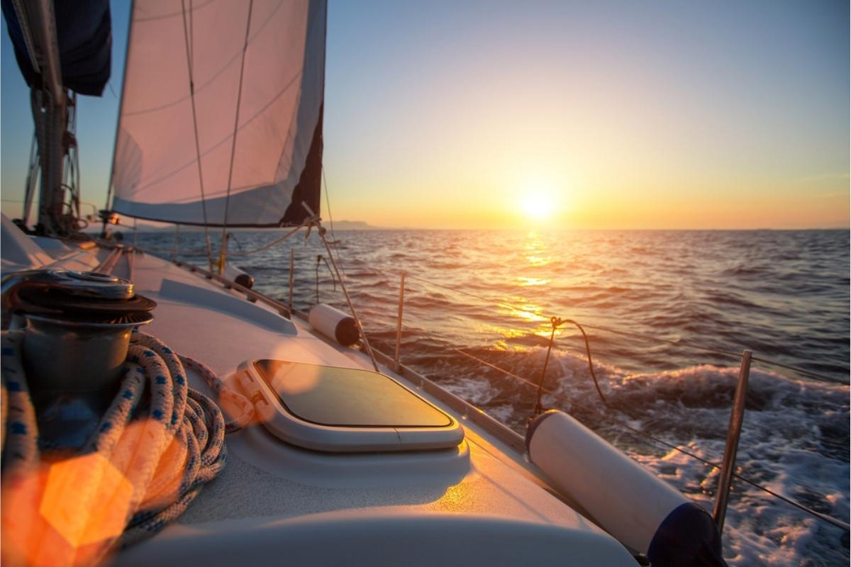 POV from a sailing yacht out on the sea at sunset. The water is blue and grey, but reflected the orange of the setting sun. You can see part of the white boat in front of you, including the sail.