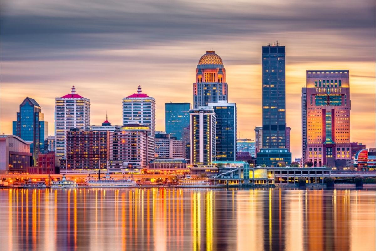 Louisville skyline and the Ohio River at sunset with tall and medium-sized buildings in the background, river in the foreground, and orange, yellow, and gold light in the sky and reflected on the water.