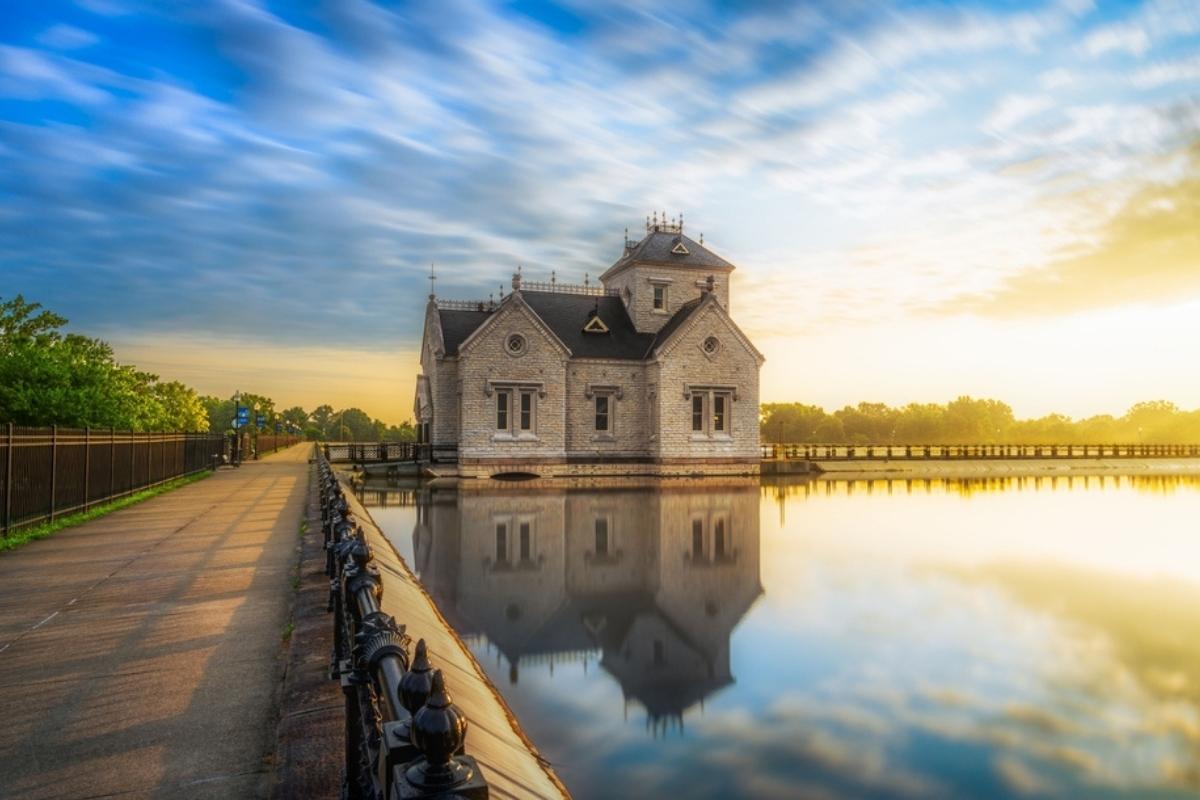 Gothic-style Louisville Reservoir in early morning sunlight. Building is beside a wide walking path along the water with iron fencing on the left side of the path, green trees around the edge, and blue sky overhead. The sunlight is shining down from the right, reflecting building and sky in the reservoir water.