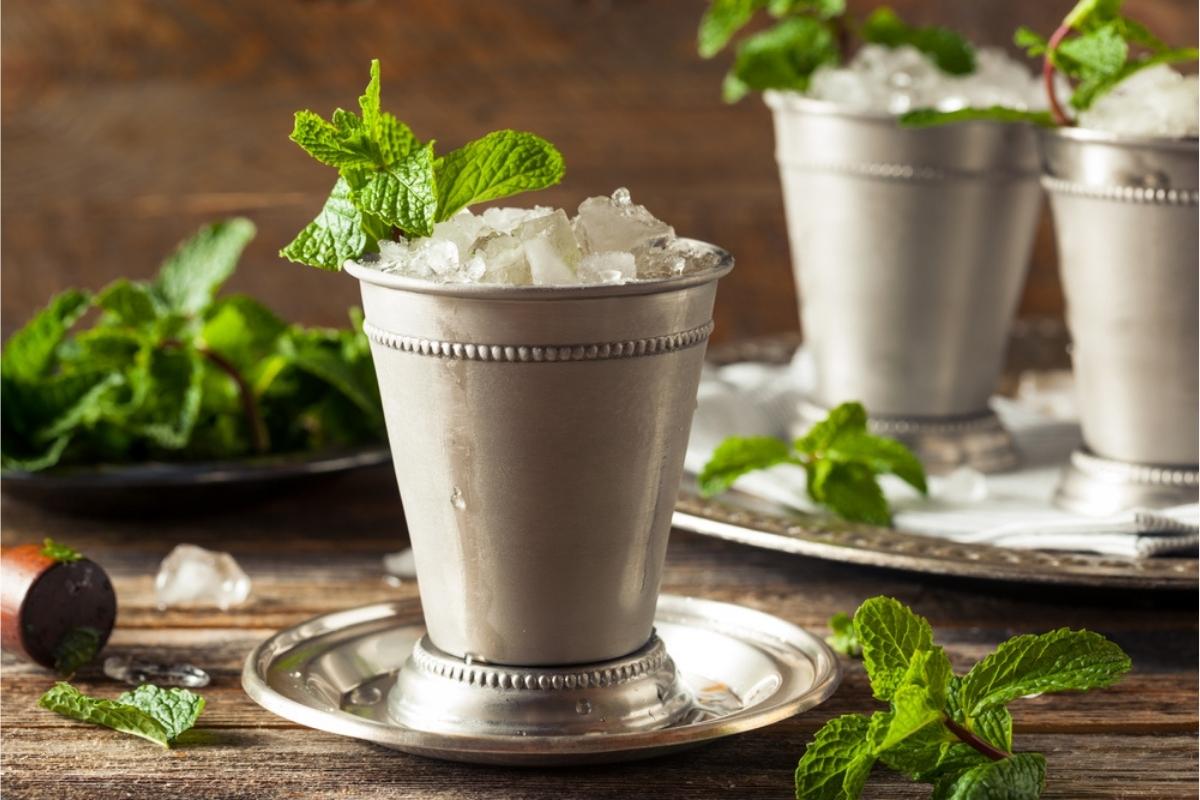 Three ice cold mint juleps in traditional mint julep cups on a silver tray set on a wooden table with mint leaves placed beside them.