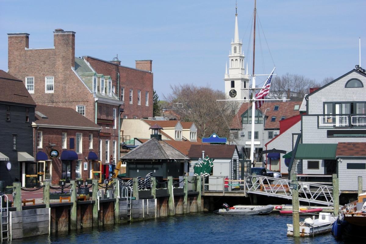 The waterfront in old town Newport in fall, with small empty boats on blue water, tied up at the dock, rows of old brick and wooden buildings, and a tall white steeple in the background.