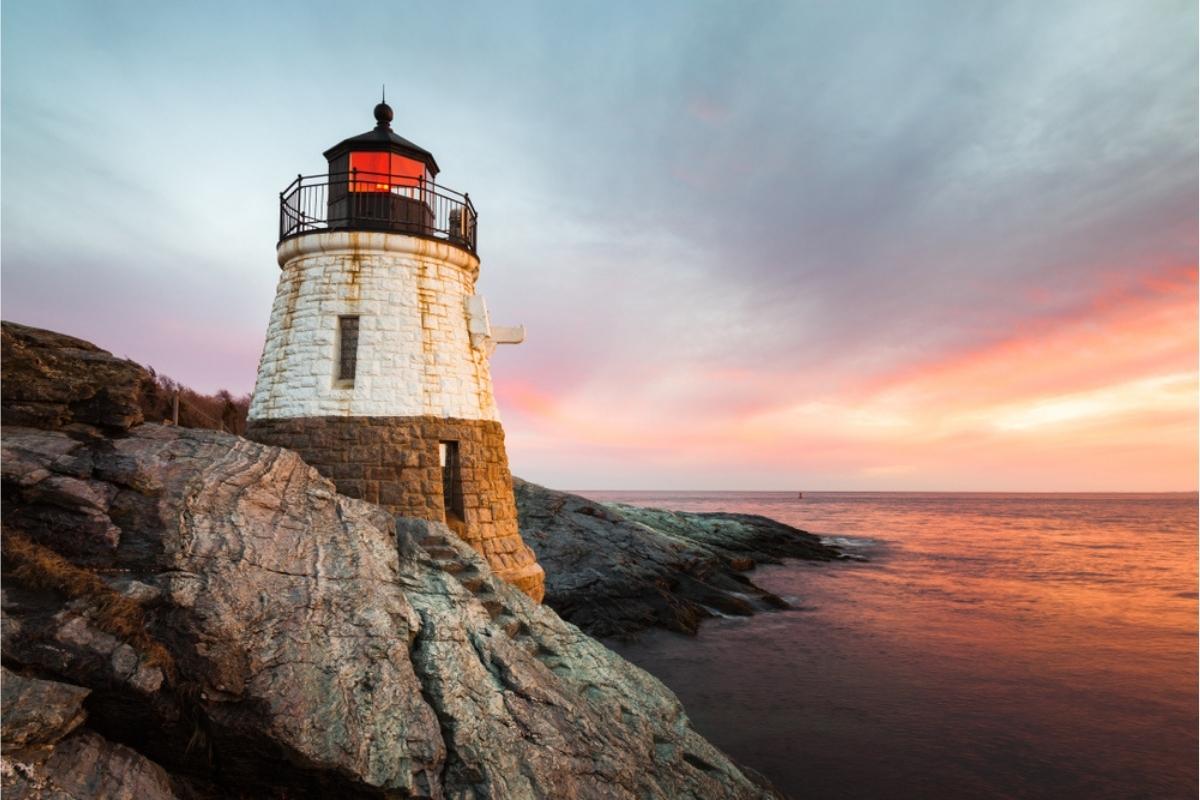 Old New England lighthouse at sunset. Lighthouse is brown and white stone with railing and light at top, set on rocky coastline, with the pink, blue, and orange sky reflected in the water below.