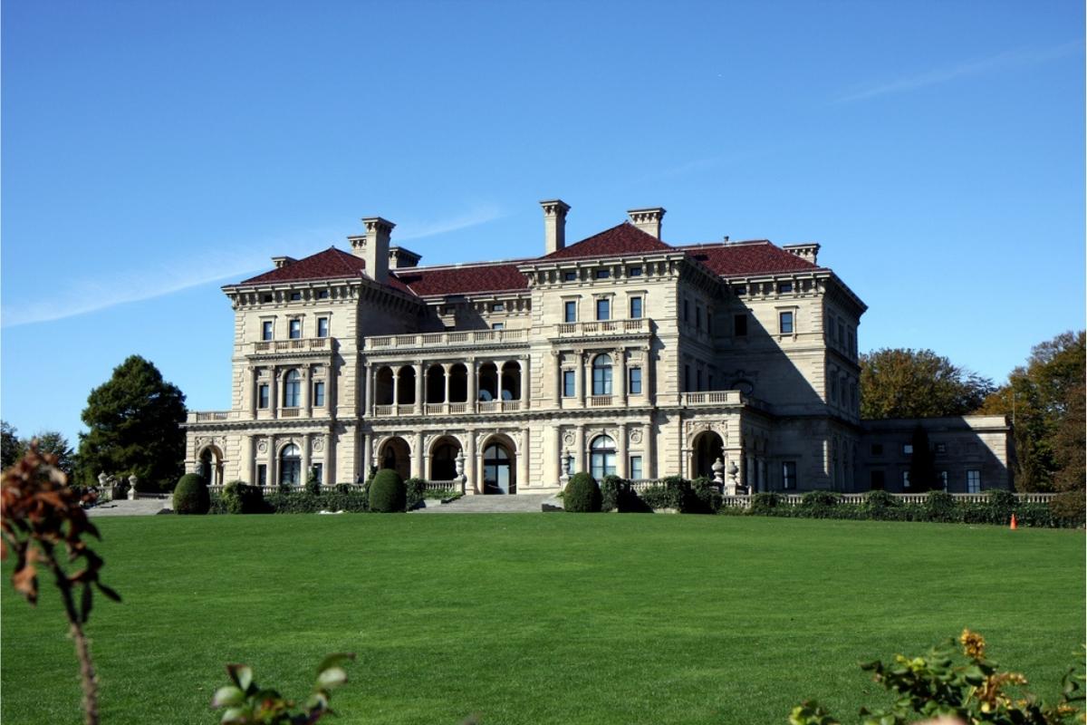 Ornate Gilded Age mansion in Newport, Rhode Island. The mansion is set on a manicured great lawn with trees and hedges along the side and in the back and deep blue sky overhead.