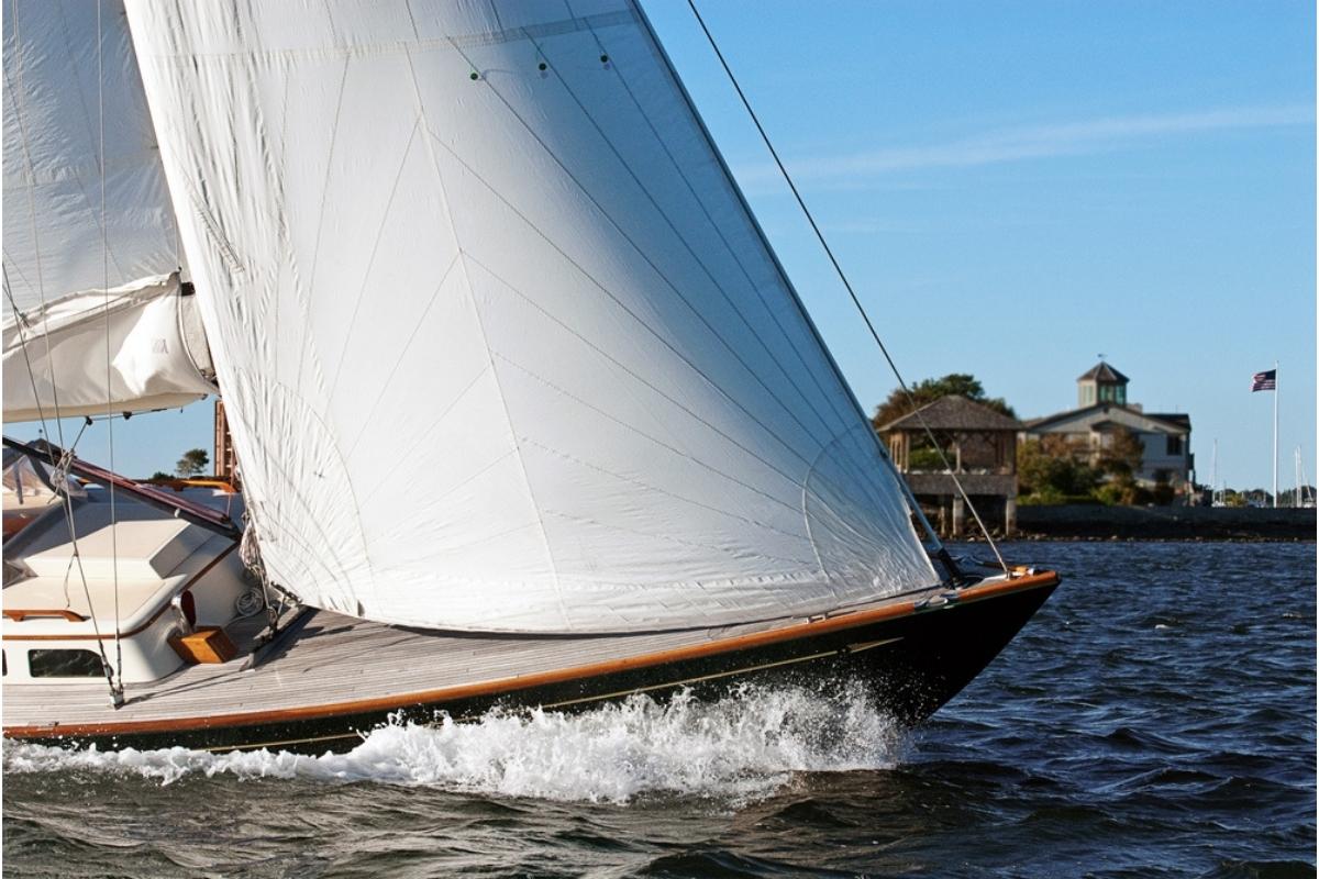 Close up shot of a sailboat on the choppy blue water in Newport Harbor.  Sails are full, the sky is blue, and there's a building with a flag flying in the background.