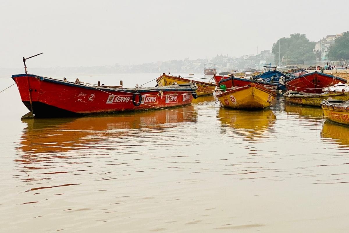 Traditional red, yellow, and blue Indian boats on the brown water of the Ganges River. The boats are empty and tied to shore. In the hazy background, there are trees, buildings, and people walking along the water. The sky above is hazy and grey.