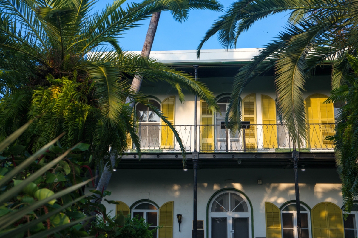 Exterior view of Hemingway House in Key West, with yellow shutters, lush green landscaping and tall palm trees, and blue sky overhead