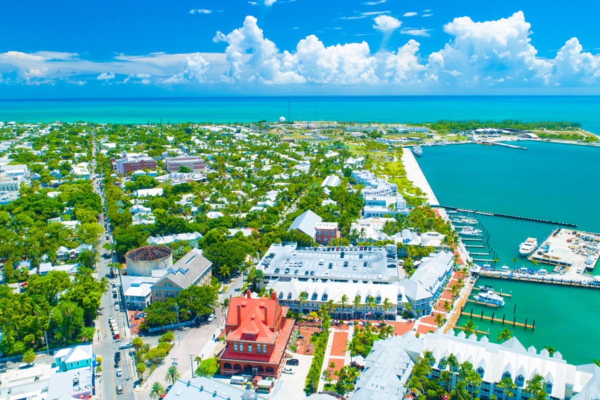 Aerial view of Key West with small marina, retail and residential streets, and green trees and landscaping. The water is blue green and the sky above is a darker blue with white, fluffy clouds