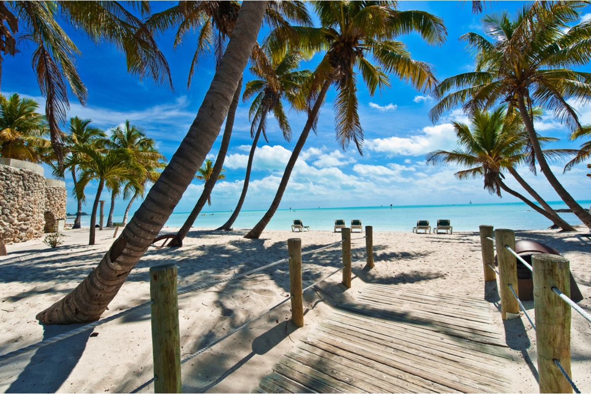 Key West beach with leaning palm trees, a low wooden pathway leading to the sand, a few beach chairs set up by the water and dark blue sky with white clouds overhead