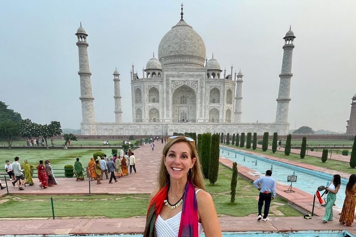 Melissa, the blog post author, smiling as she stands in front of the walkway leading to the Taj Mahal, groups of people, grass, water, and trees are in the background. The grey and white stone structure of The Taj Mahal fills the background and the sky is grey and blue overhead.