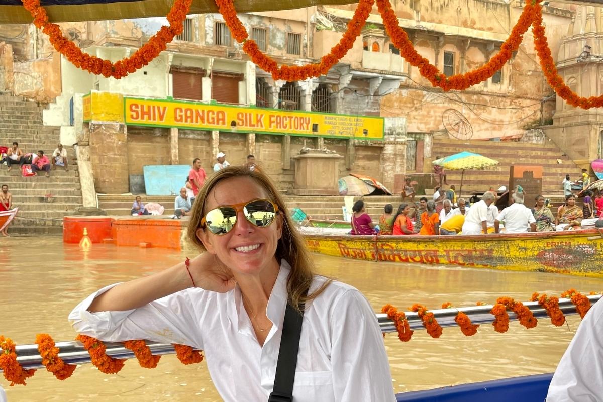 Melissa, the blog post author, smiling and wearing white clothes, sunglasses, and a red string around her wrist, riding in a traditional Indian boat on the brown water of the Ganges River. Other boats with people are in the background.