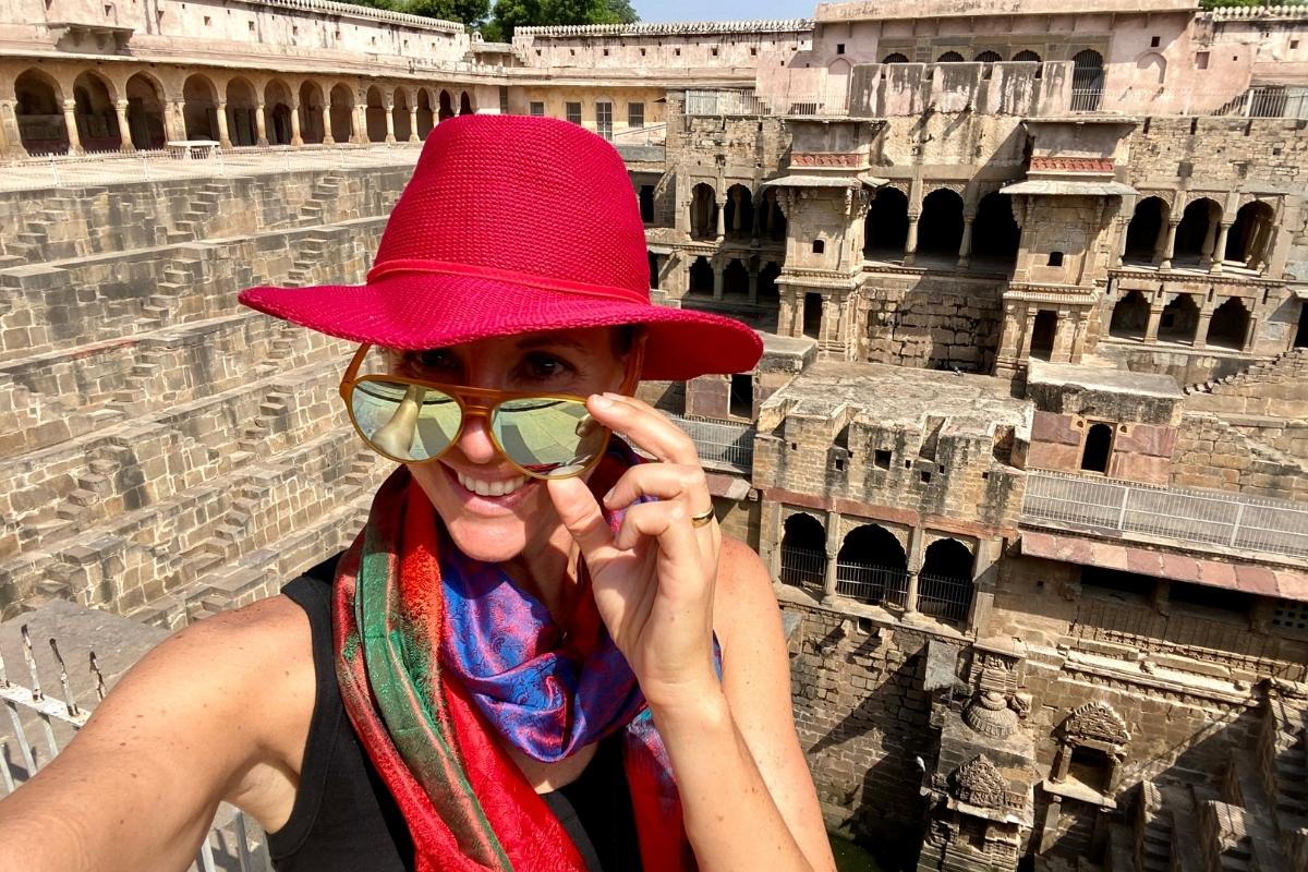 Melissa, the blog post author, smiling and wearing a reddish-pink sun hat, bright scarf, black top and sunglasses, taking a selfie in front of a towering stone stepwell on the way to Agra, India.