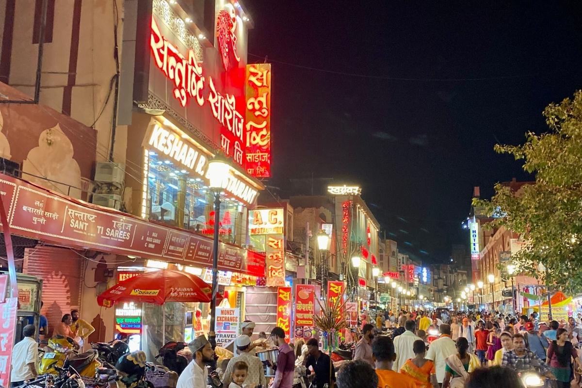 Busy Varanasi street at night with well-lit buildings and lamp posts lining each side and hundreds of people walking or riding motorbikes.