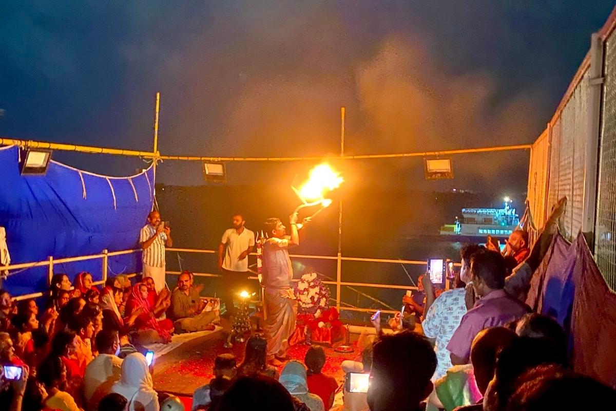 Man holding aloft a flame of fire at night as a group of people sit and stand around him on a small platform next to the Ganges River. A wooden frame surrounds the platform and a large dark blue piece of fabric is tied to the frame on the left side of the image.