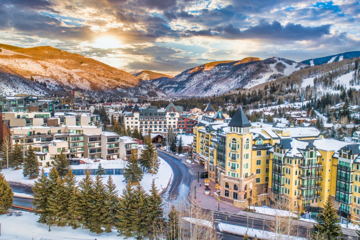 Aerial view of Vail with large resort buildings, streets lined with evergreens, and snowy mountains in the background. The sky is blue with white clouds and golden sunlight peeking through.