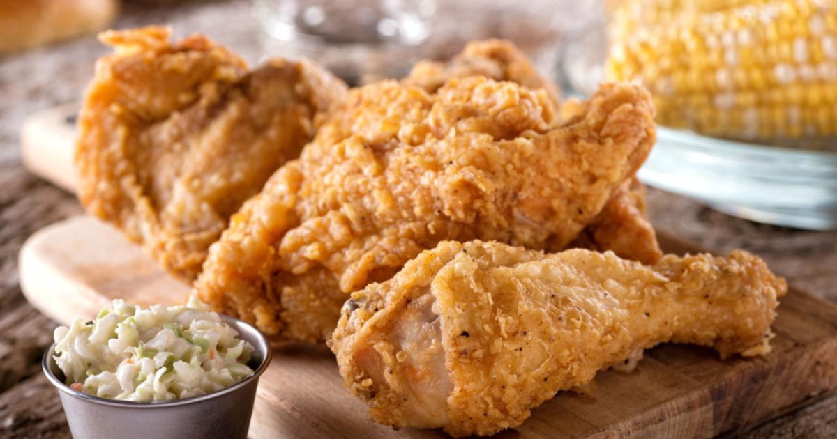 Close up shot of three pieces of fried chicken, a small side of coleslaw, and corn on the cob, set on a wooden table