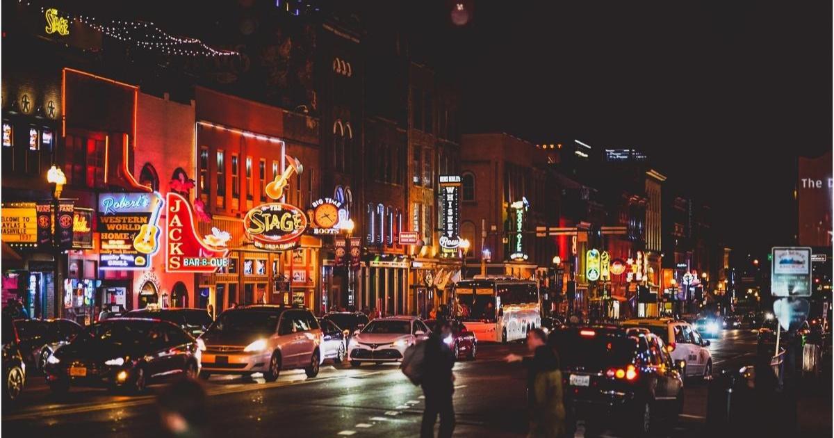 Busy nighttime street in downtown Nashville with cars, bars, pedestrians, and neon lights and signs