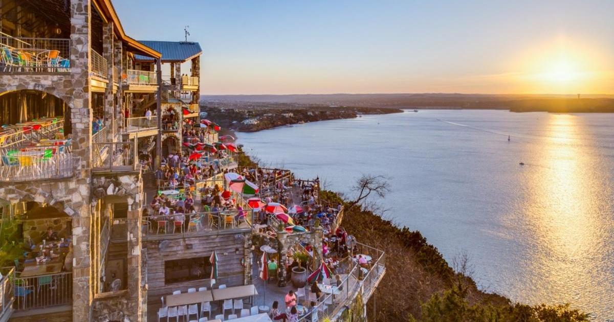 Aerial view of The Oasis at Lake Travis, with color chairs and umbrellas on the patios, tree-lined ground next to the lake below and sun setting in the distance