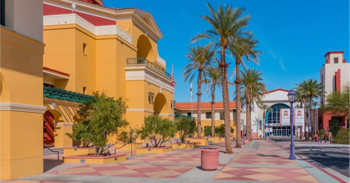 Cathedral City city center with golden yellow and white buildings, a wide walkway lined with palm trees and small shrubs, and a clear blue sky overhead