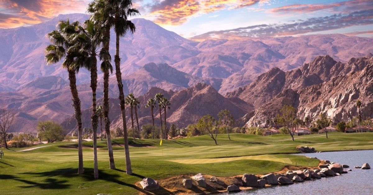 Palm Springs golf course with rolling greens alongside a small pond, a few tall palm trees, and mountains in the background