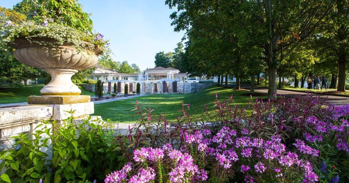 Green grass, purple and pink flowers, and lush trees in front of and alongside a white stone wall and walkways. In the background is a white building and sky overhead is a clear, light blue.